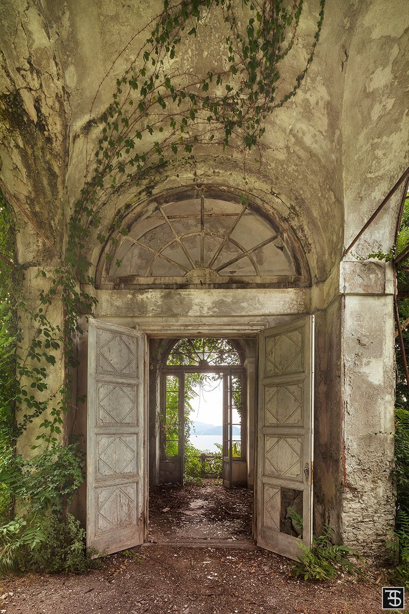 Overgrown archway with an open door revealing water beyond, Italy