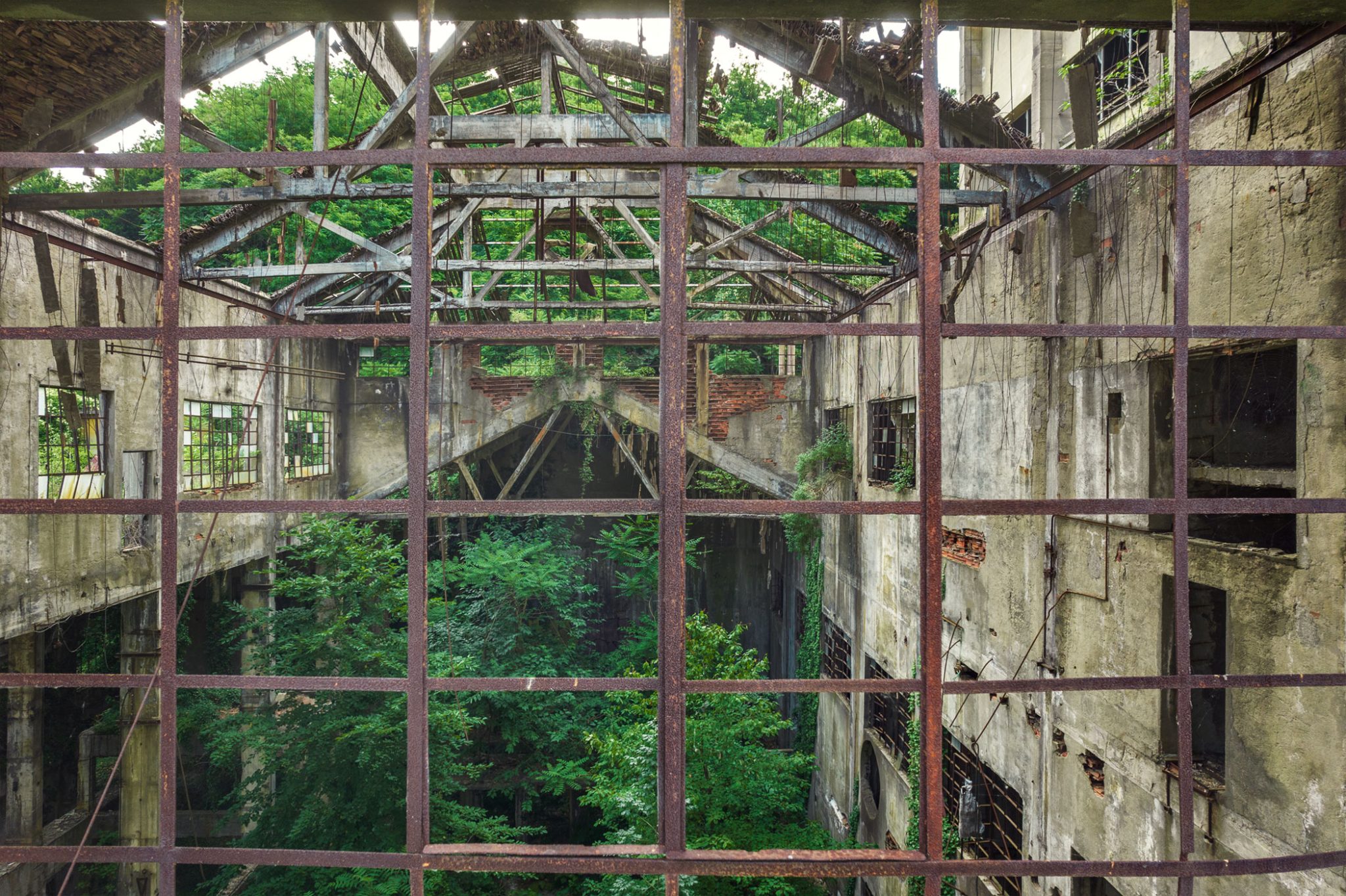 View through a rusted grid window onto a multi-storey factory ruin in Italy