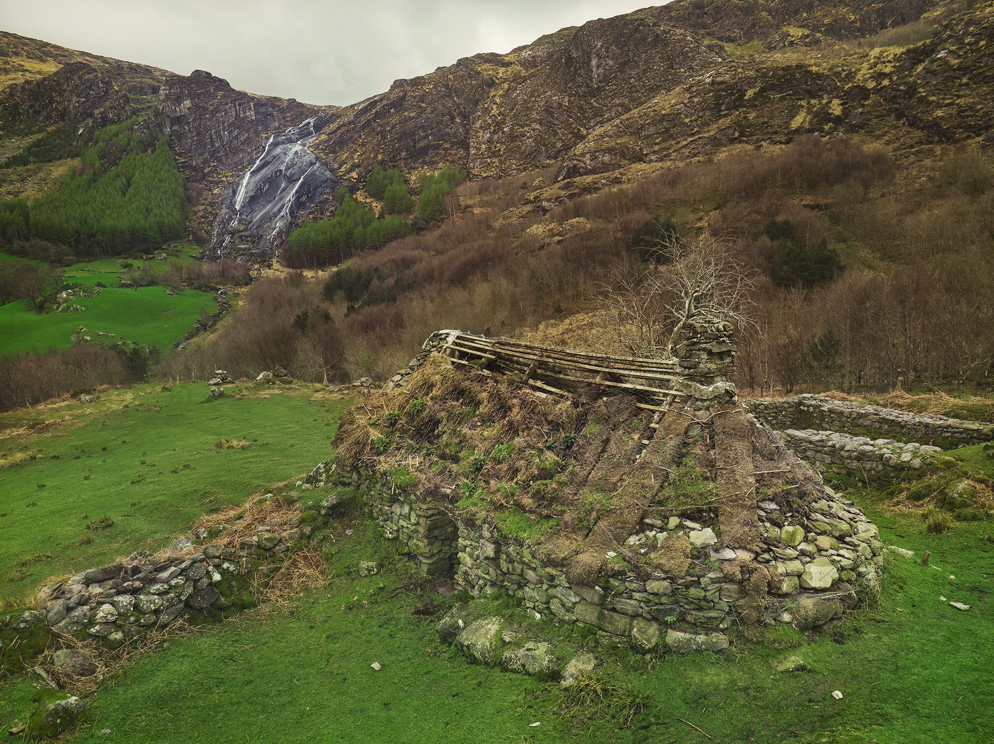 Steinhütte mit bemoostem Dach am Fuß eines Wasserfalls, Irland