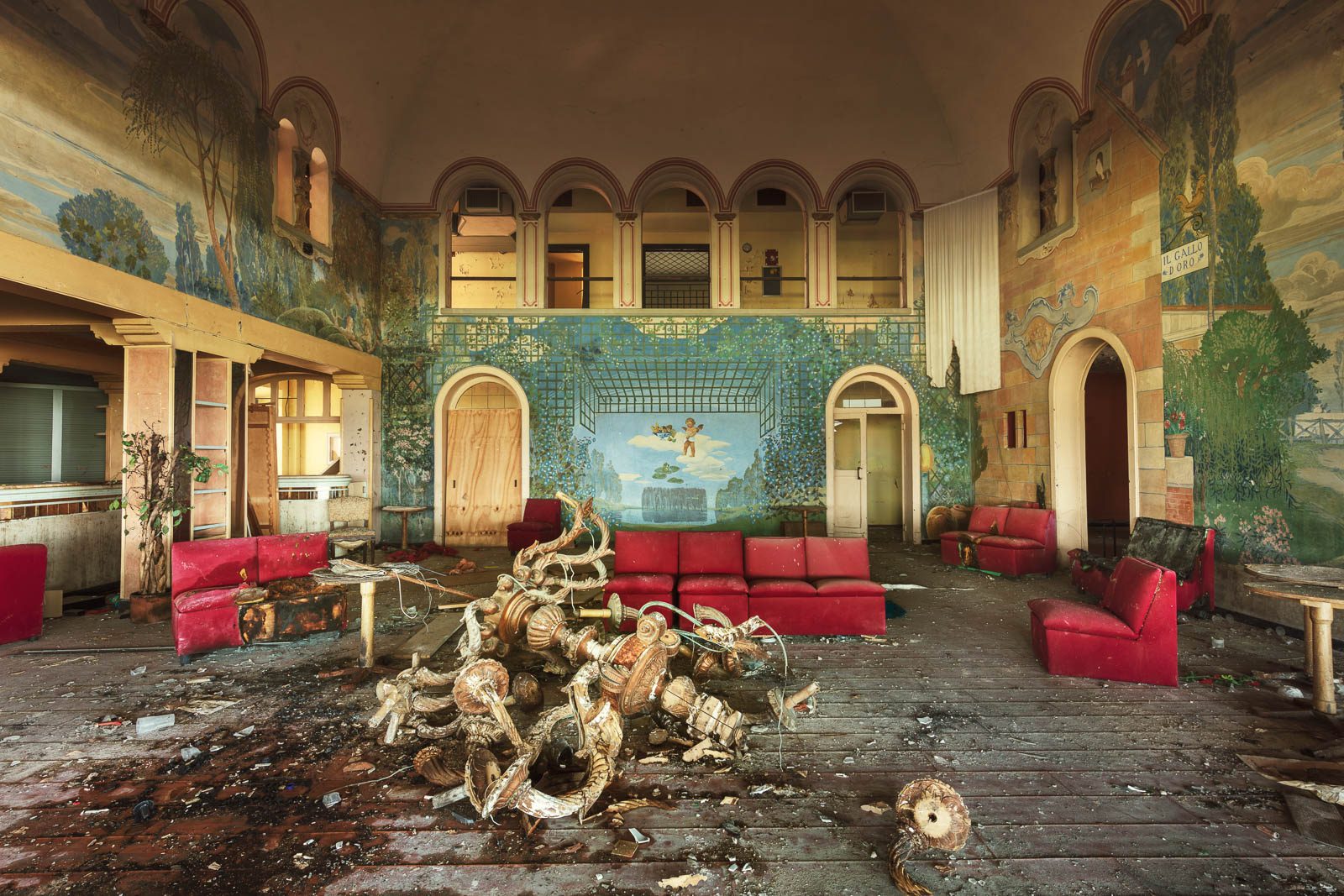 Saal mit Wandmalereien, roten Ledersofas und zerbrochenem Kronleuchter in einer ehemaligen Discothek in Italien