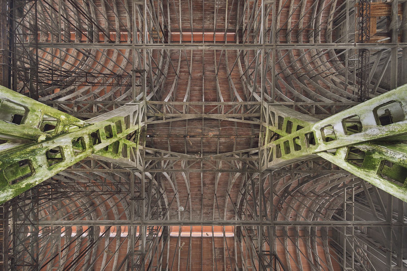 A brick-vaulted hall roof with x-shaped, moss-covered steel struts in an industrial ruin in Italy